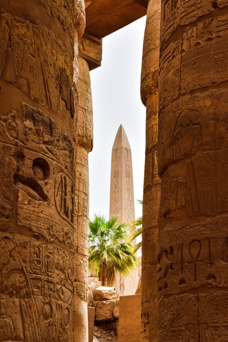 View of an Egyptian obelisk framed by ancient hieroglyph-covered pillars, with palm trees in the background.